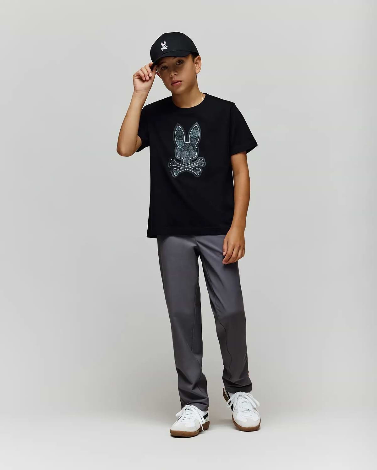 A young boy wears the Psycho Bunny KIDS ROWEN BUNNY GRAPHIC TEE (KT0200058) in black, paired with a black cap, gray pants, and white sneakers. He stands against a light gray background, holding the brim of his cap. (4)
