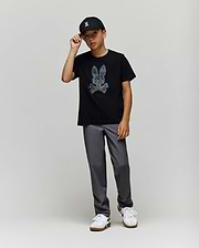 A young boy wears the Psycho Bunny KIDS ROWEN BUNNY GRAPHIC TEE (KT0200058) in black, paired with a black cap, gray pants, and white sneakers. He stands against a light gray background, holding the brim of his cap. (4)
