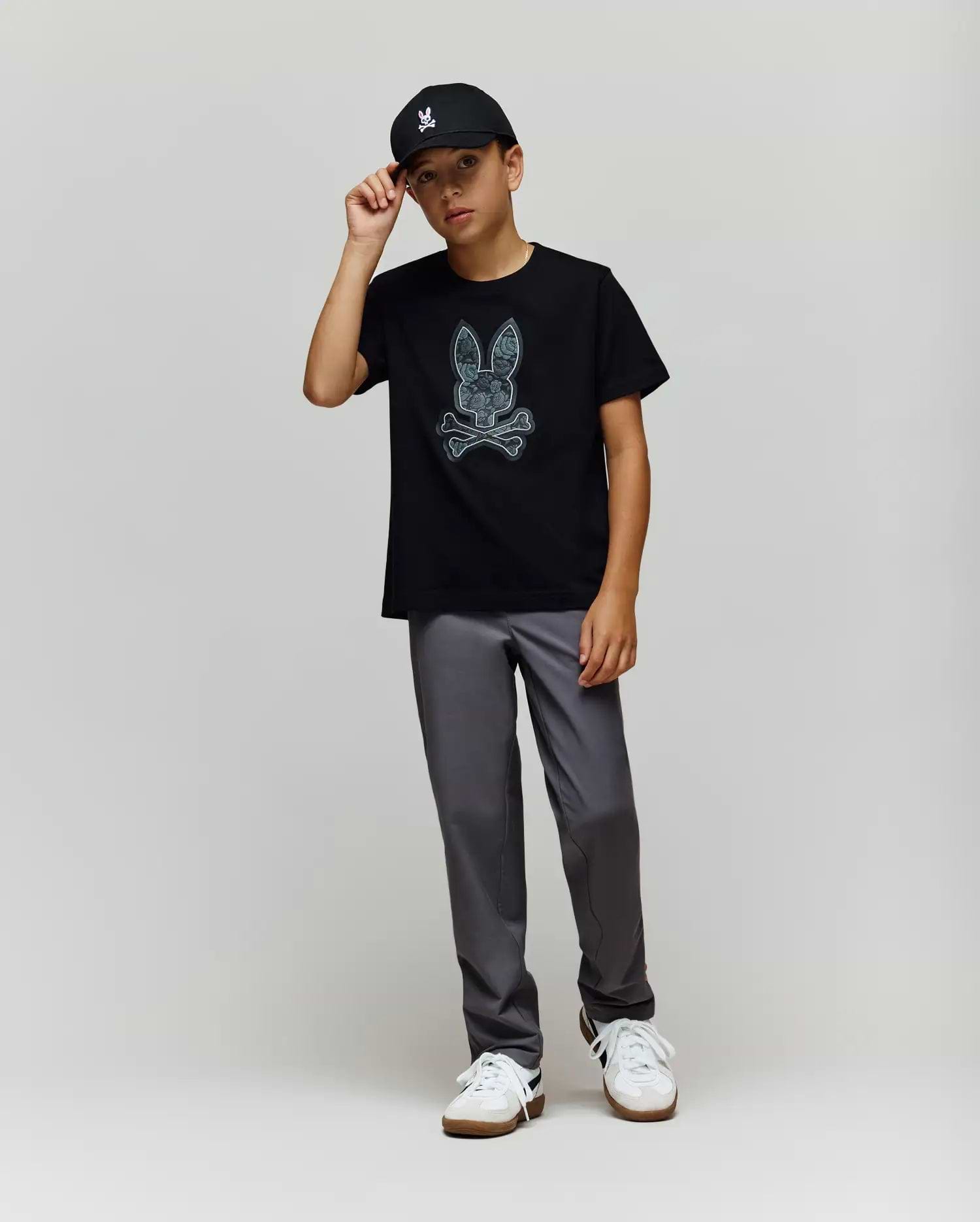 A young boy wears the Psycho Bunny KIDS ROWEN BUNNY GRAPHIC TEE (KT0200058) in black, paired with a black cap, gray pants, and white sneakers. He stands against a light gray background, holding the brim of his cap. (4)