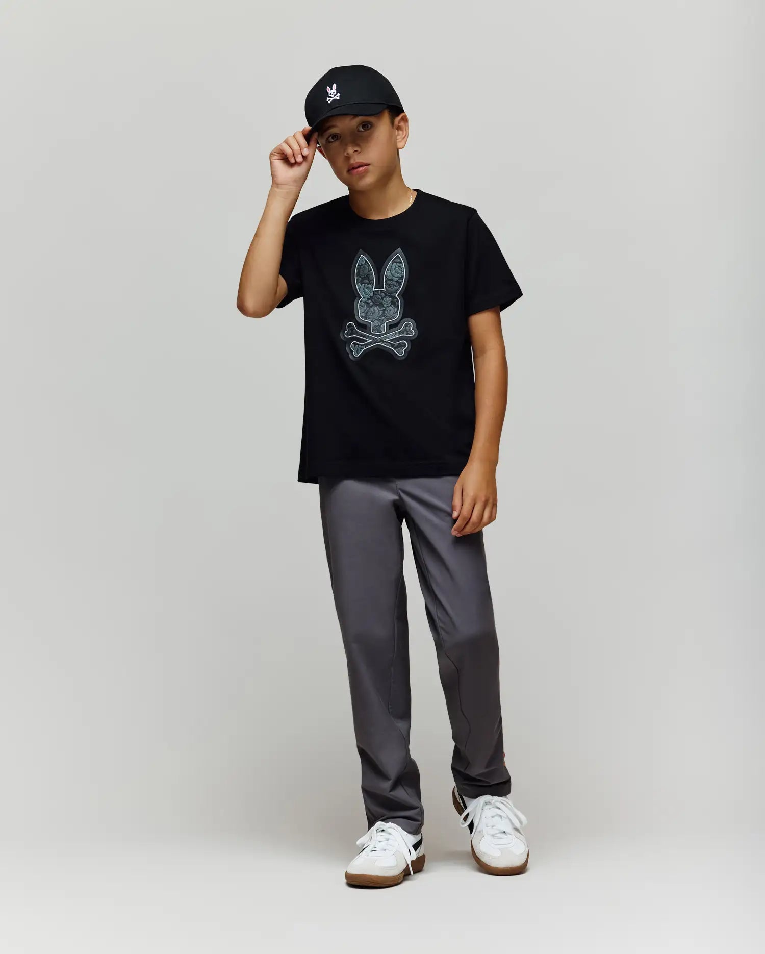 A young boy wears the Psycho Bunny KIDS ROWEN BUNNY GRAPHIC TEE (KT0200058) in black, paired with a black cap, gray pants, and white sneakers. He stands against a light gray background, holding the brim of his cap. (4)