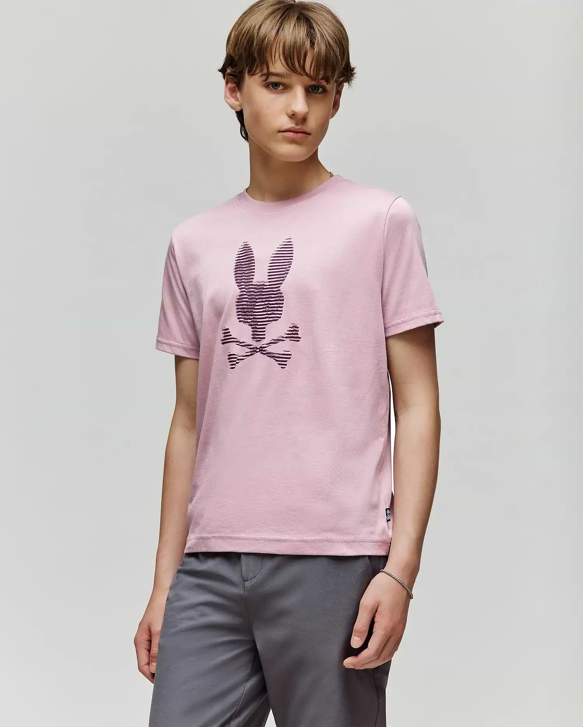 A young person with light brown hair wears the Psycho Bunny KIDS GRANT BUNNY GRAPHIC TEE (KT0200061) in light pink Pima cotton, featuring an HD-printed bunny head above crossed bones, paired with gray shorts against a plain background.