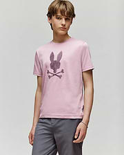 A young person with light brown hair wears the Psycho Bunny KIDS GRANT BUNNY GRAPHIC TEE (KT0200061) in light pink Pima cotton, featuring an HD-printed bunny head above crossed bones, paired with gray shorts against a plain background.