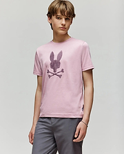 A young person with light brown hair wears the Psycho Bunny KIDS GRANT BUNNY GRAPHIC TEE (KT0200061) in light pink Pima cotton, featuring an HD-printed bunny head above crossed bones, paired with gray shorts against a plain background.