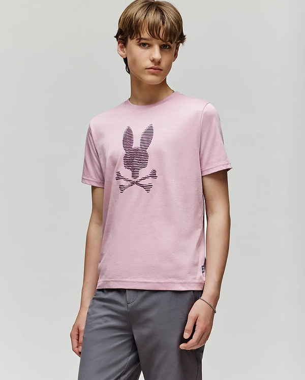 A young person with light brown hair wears the Psycho Bunny KIDS GRANT BUNNY GRAPHIC TEE (KT0200061) in light pink Pima cotton, featuring an HD-printed bunny head above crossed bones, paired with gray shorts against a plain background.