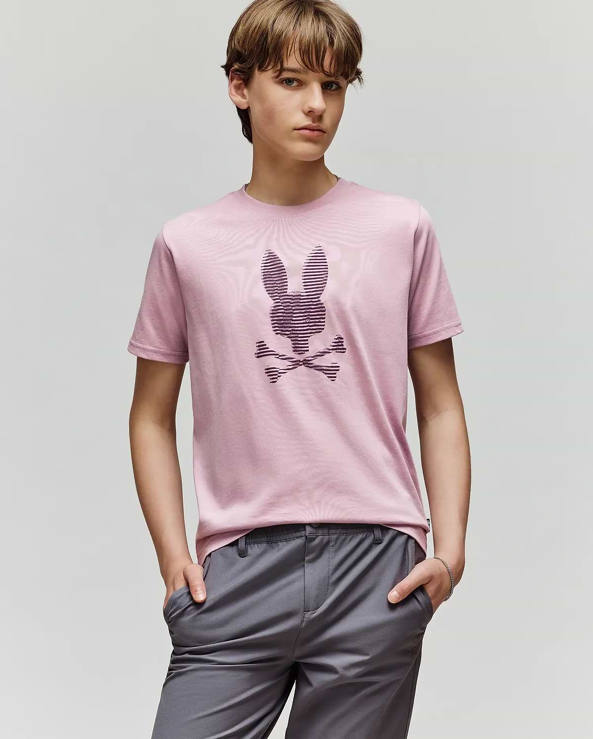 A young person with light brown hair wears the Psycho Bunny KIDS GRANT BUNNY GRAPHIC TEE (KT0200061) in light pink, featuring a stylized bunny with crossbones, paired with gray pants and standing hands-in-pockets against a plain background.
