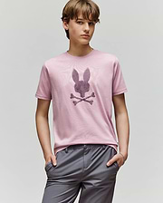 A young person with light brown hair wears the Psycho Bunny KIDS GRANT BUNNY GRAPHIC TEE (KT0200061) in light pink, featuring a stylized bunny with crossbones, paired with gray pants and standing hands-in-pockets against a plain background.