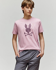 A young person with light brown hair wears the Psycho Bunny KIDS GRANT BUNNY GRAPHIC TEE (KT0200061) in light pink, featuring a stylized bunny with crossbones, paired with gray pants and standing hands-in-pockets against a plain background.