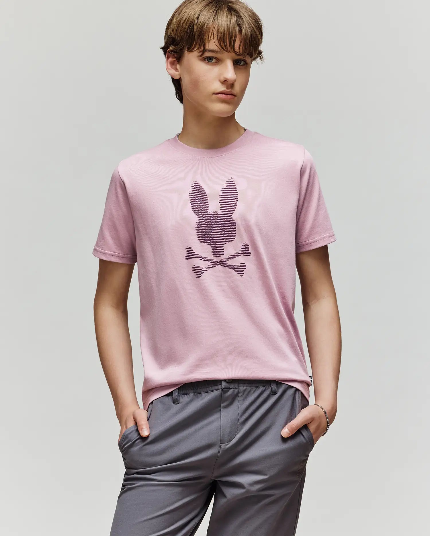 A young person with light brown hair wears the Psycho Bunny KIDS GRANT BUNNY GRAPHIC TEE (KT0200061) in light pink, featuring a stylized bunny with crossbones, paired with gray pants and standing hands-in-pockets against a plain background.