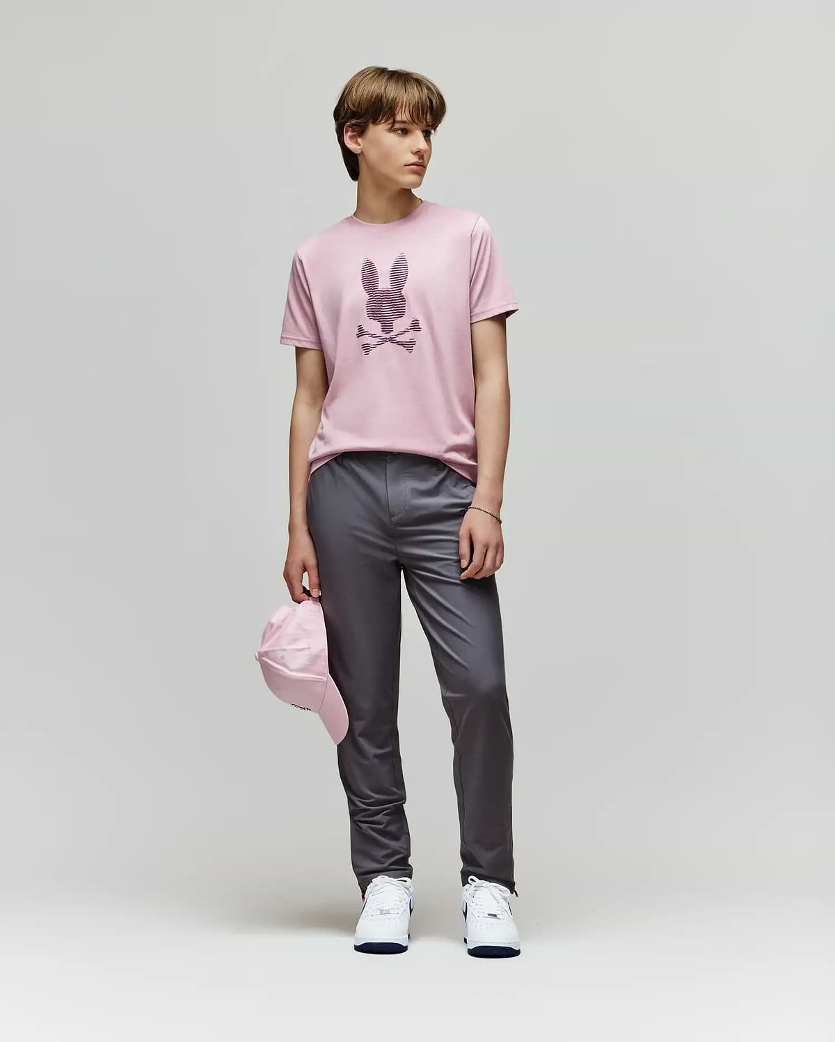 A person stands against a plain background wearing the Psycho Bunny KIDS GRANT BUNNY GRAPHIC TEE - KT0200061, gray pants, white sneakers, and holding a pink cap by their side.