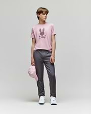 A person stands against a plain background wearing the Psycho Bunny KIDS GRANT BUNNY GRAPHIC TEE - KT0200061, gray pants, white sneakers, and holding a pink cap by their side.