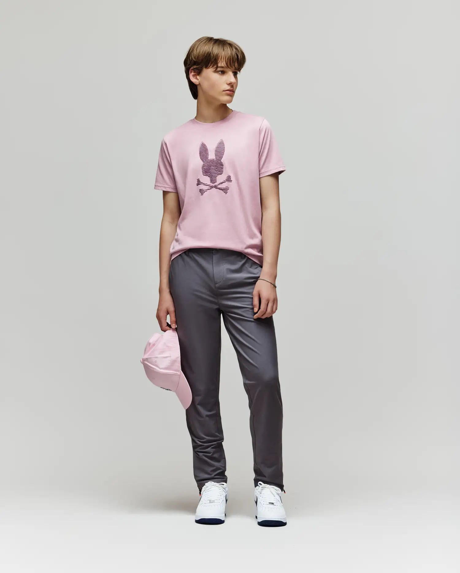 A person stands against a plain background wearing the Psycho Bunny KIDS GRANT BUNNY GRAPHIC TEE - KT0200061, gray pants, white sneakers, and holding a pink cap by their side.