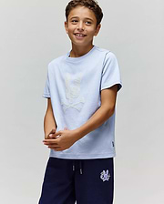 A smiling boy wears the Psycho Bunny KIDS GRANT BUNNY GRAPHIC TEE (KT0200061), HD-printed on soft Pima cotton with a white bunny and crossbones, paired with navy shorts, standing against a light background. (2)
