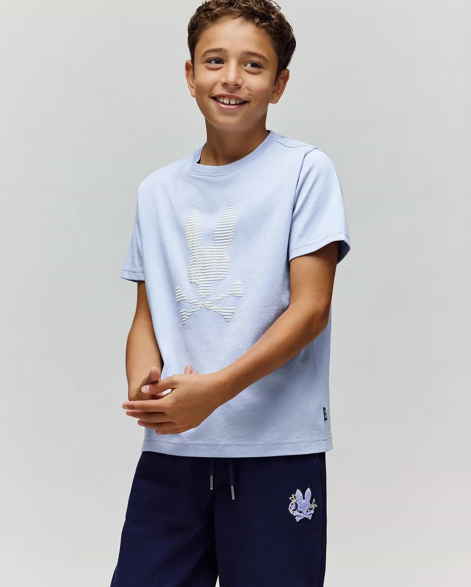 A smiling boy wears the Psycho Bunny KIDS GRANT BUNNY GRAPHIC TEE (KT0200061), HD-printed on soft Pima cotton with a white bunny and crossbones, paired with navy shorts, standing against a light background. (2)