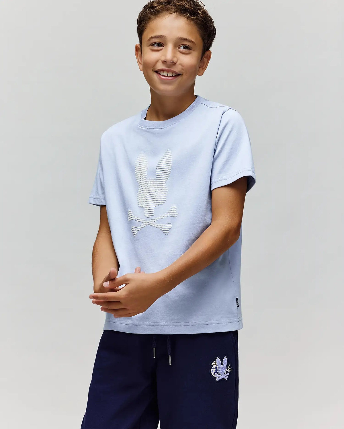 A smiling boy wears the Psycho Bunny KIDS GRANT BUNNY GRAPHIC TEE (KT0200061), HD-printed on soft Pima cotton with a white bunny and crossbones, paired with navy shorts, standing against a light background. (2)