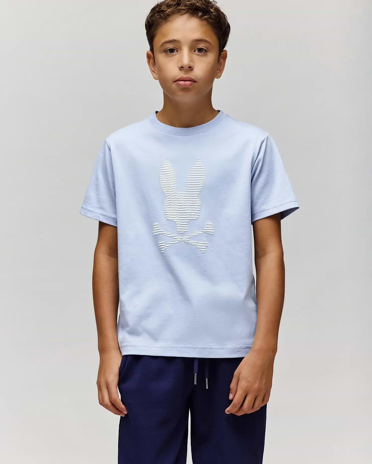 A boy stands against a plain light gray background, wearing the Psycho Bunny KIDS GRANT BUNNY GRAPHIC TEE (KT0200061) in light blue with a white skull and crossbones bunny graphic, paired with dark blue pants. (4)