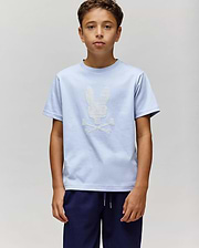 A boy stands against a plain light gray background, wearing the Psycho Bunny KIDS GRANT BUNNY GRAPHIC TEE (KT0200061) in light blue with a white skull and crossbones bunny graphic, paired with dark blue pants. (4)