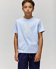 A boy stands against a plain light gray background, wearing the Psycho Bunny KIDS GRANT BUNNY GRAPHIC TEE (KT0200061) in light blue with a white skull and crossbones bunny graphic, paired with dark blue pants. (4)