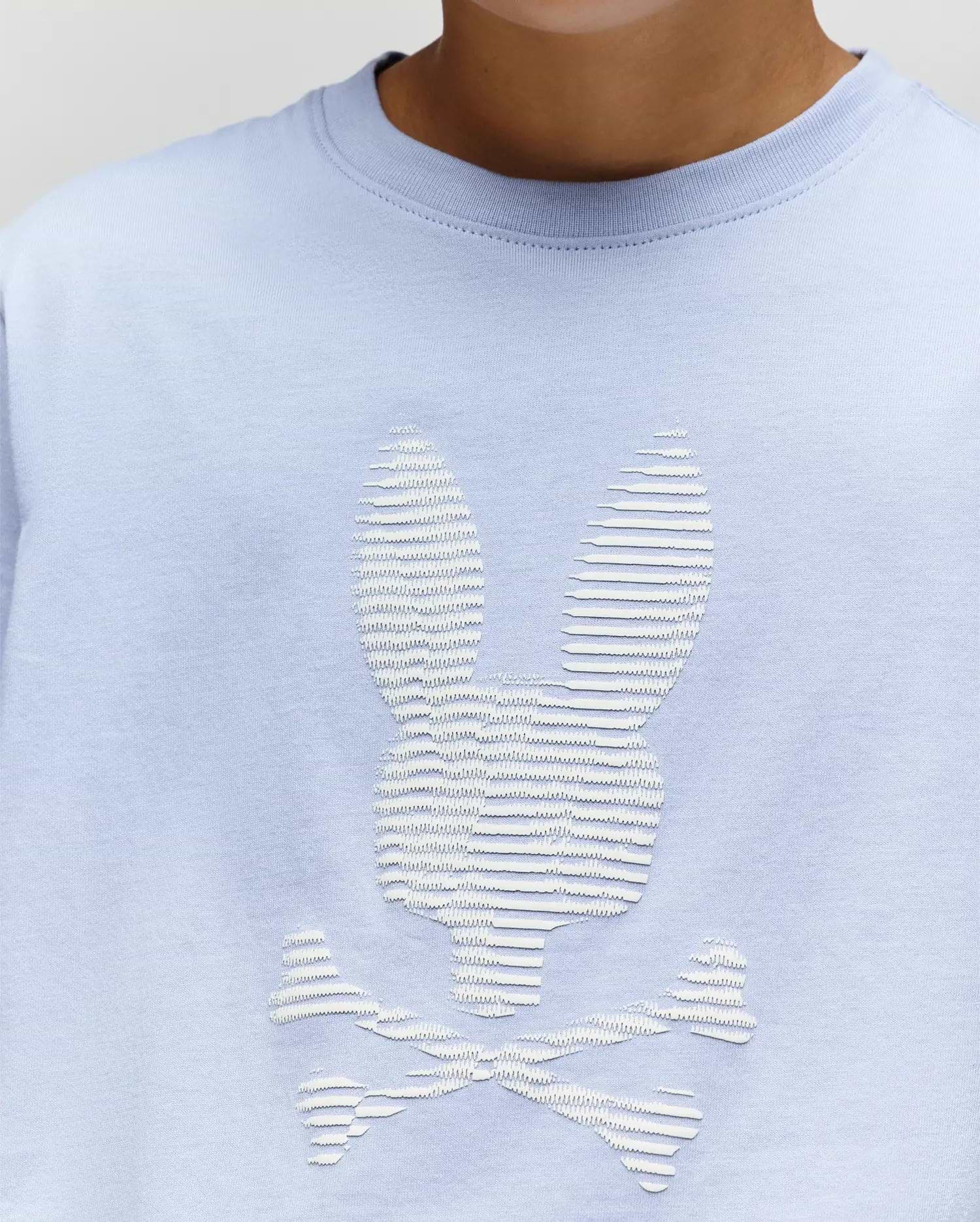 The KIDS GRANT BUNNY GRAPHIC TEE by Psycho Bunny (KT0200061) is a light blue Pima cotton shirt for kids, featuring a white textured bunny-head skull and crossbones graphic, shown worn by a child from shoulders to just below the chin. (3)