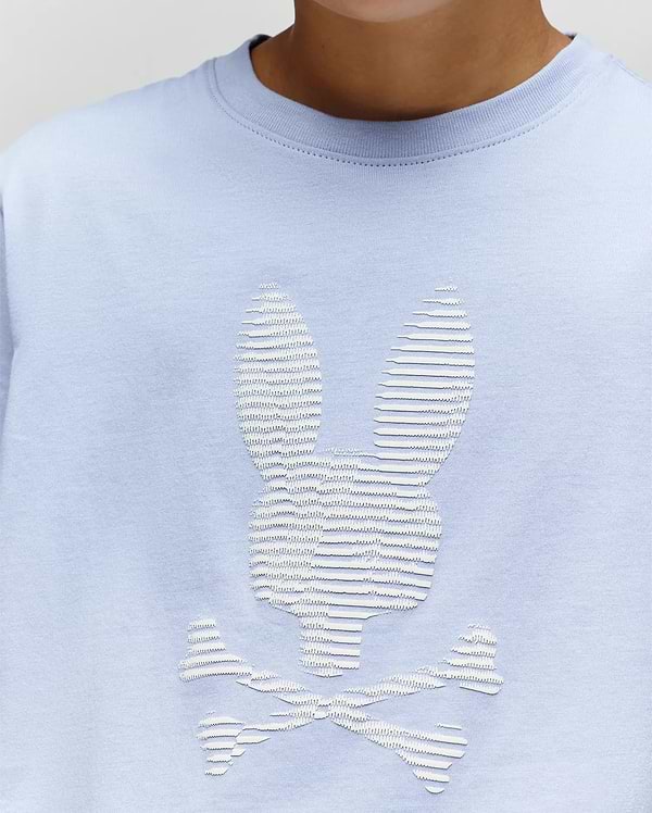 The KIDS GRANT BUNNY GRAPHIC TEE by Psycho Bunny (KT0200061) is a light blue Pima cotton shirt for kids, featuring a white textured bunny-head skull and crossbones graphic, shown worn by a child from shoulders to just below the chin. (3)