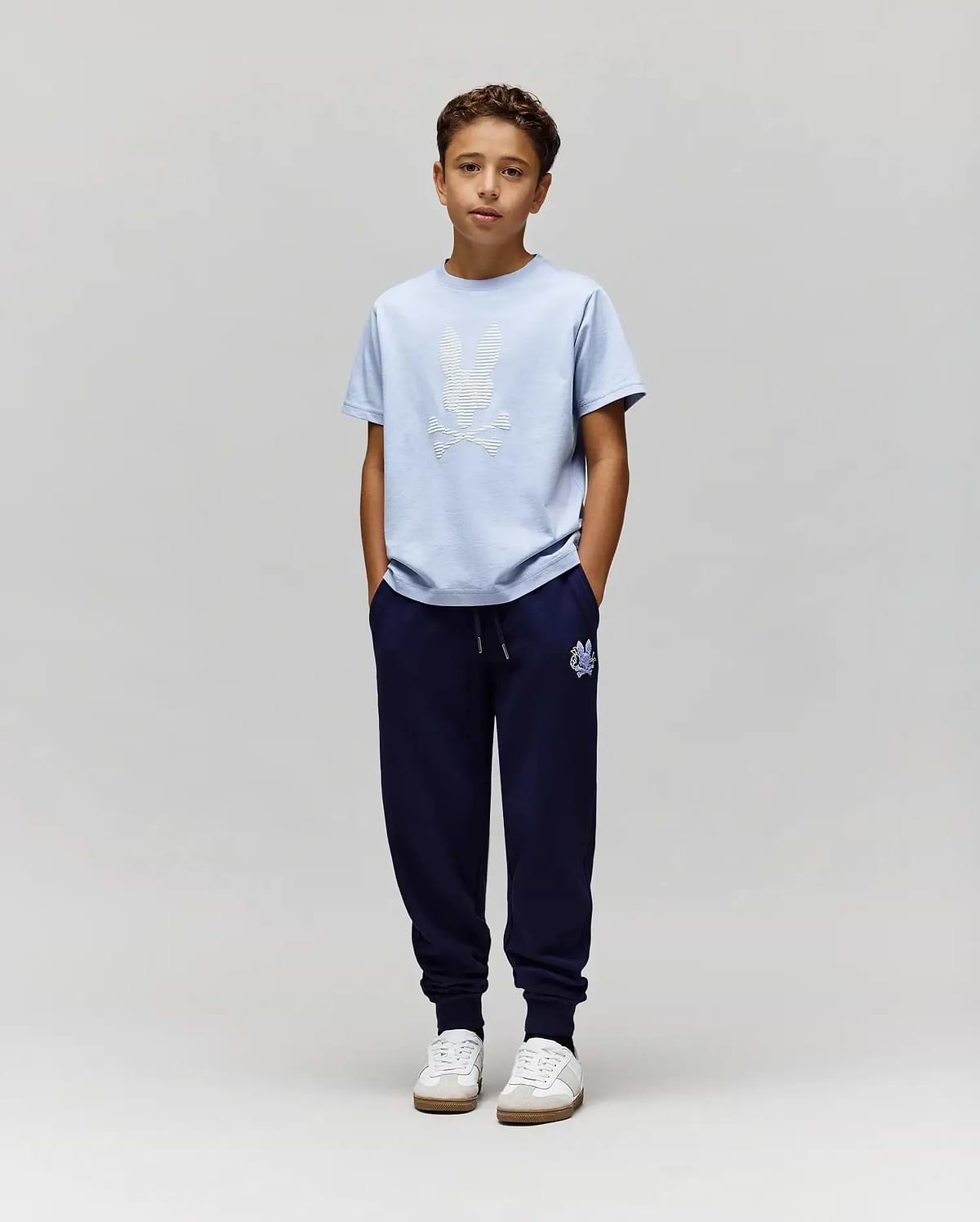 A boy with short brown hair stands against a plain light gray background, wearing Psycho Bunny's KIDS GRANT BUNNY GRAPHIC TEE (KT0200061) in light blue, dark blue jogger pants, and white sneakers with his hands in his pockets. (5)