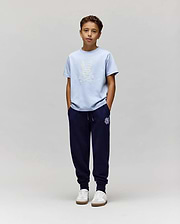 A boy with short brown hair stands against a plain light gray background, wearing Psycho Bunny's KIDS GRANT BUNNY GRAPHIC TEE (KT0200061) in light blue, dark blue jogger pants, and white sneakers with his hands in his pockets. (5)