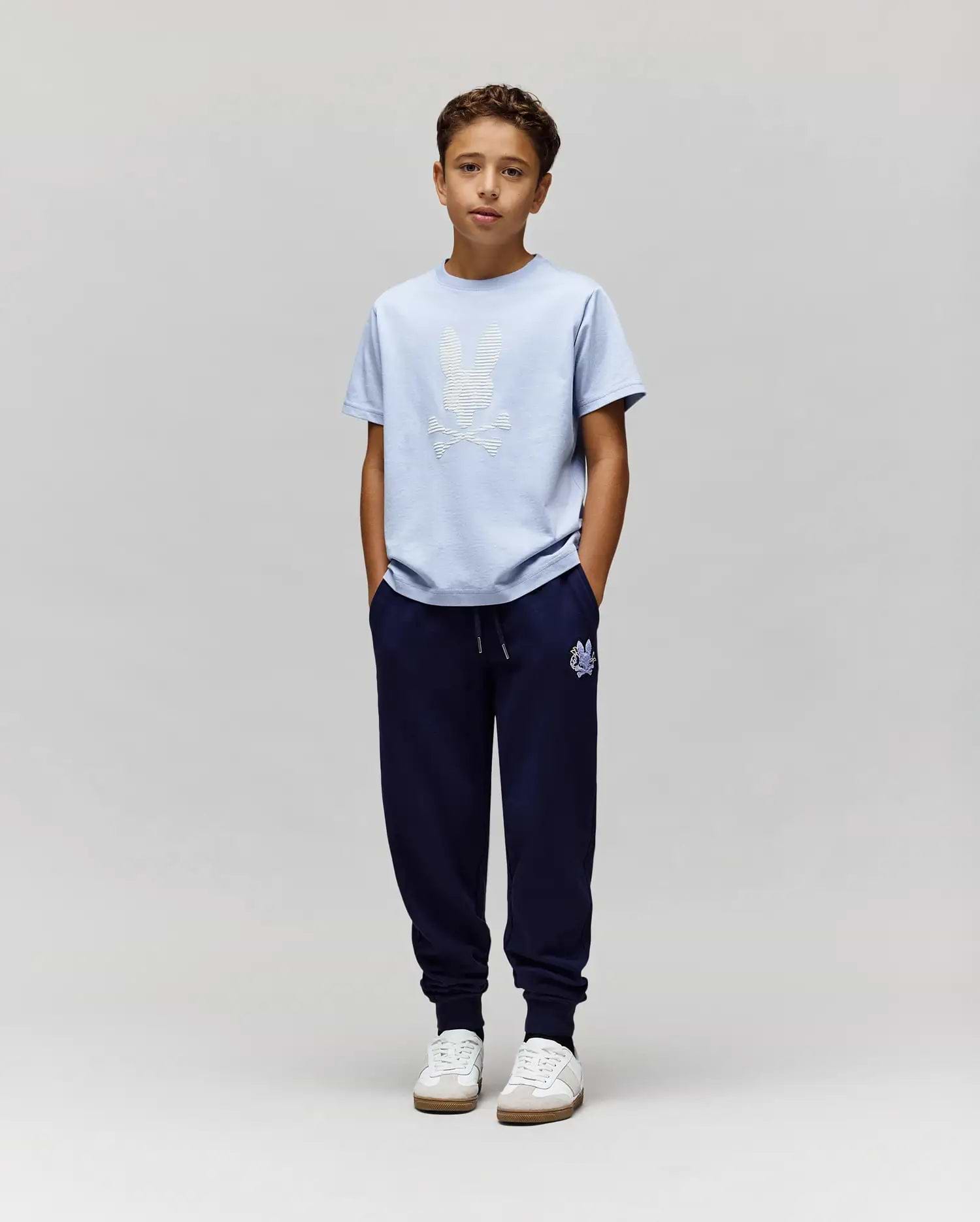 A boy with short brown hair stands against a plain light gray background, wearing Psycho Bunny's KIDS GRANT BUNNY GRAPHIC TEE (KT0200061) in light blue, dark blue jogger pants, and white sneakers with his hands in his pockets. (5)