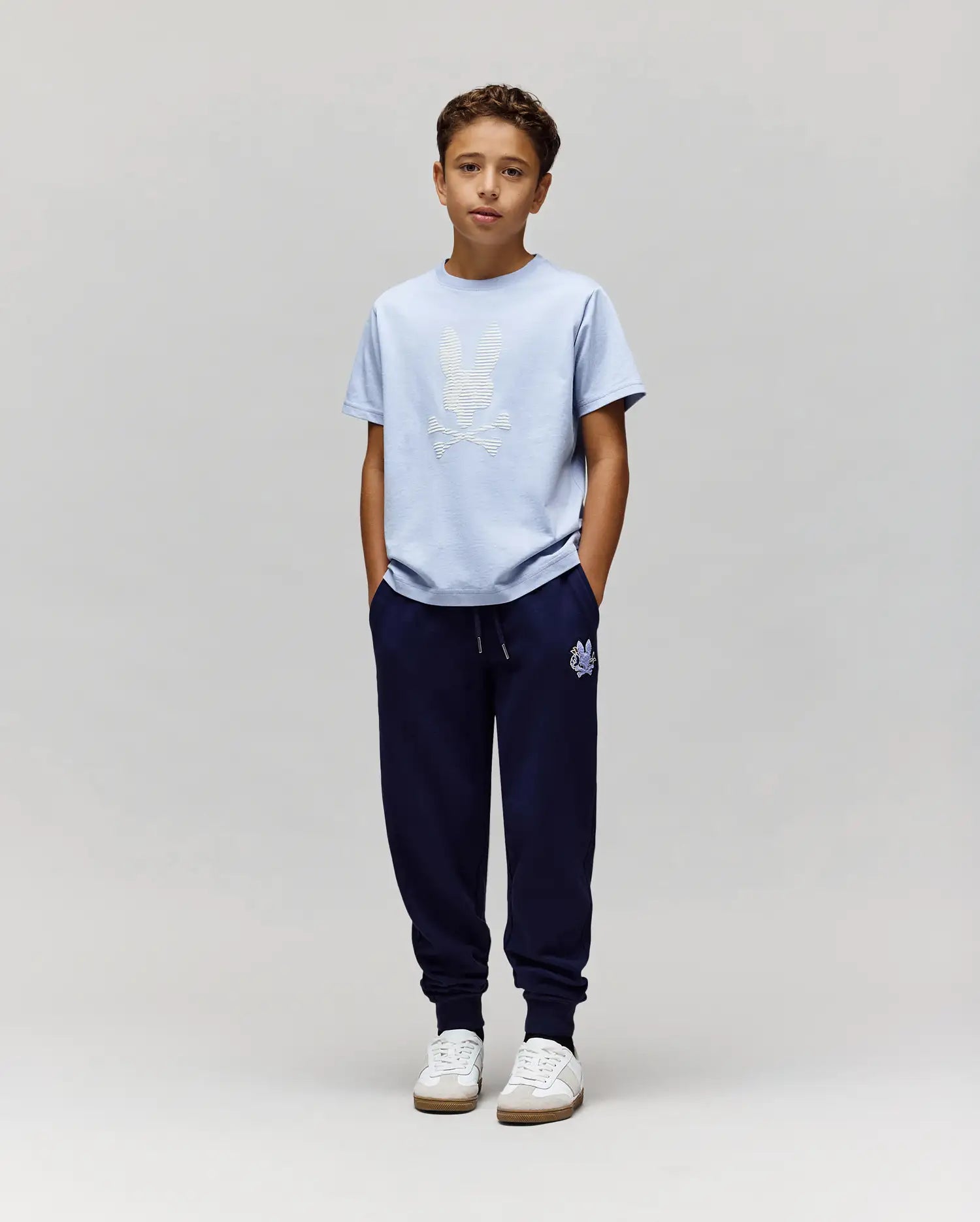 A boy with short brown hair stands against a plain light gray background, wearing Psycho Bunny's KIDS GRANT BUNNY GRAPHIC TEE (KT0200061) in light blue, dark blue jogger pants, and white sneakers with his hands in his pockets. (5)