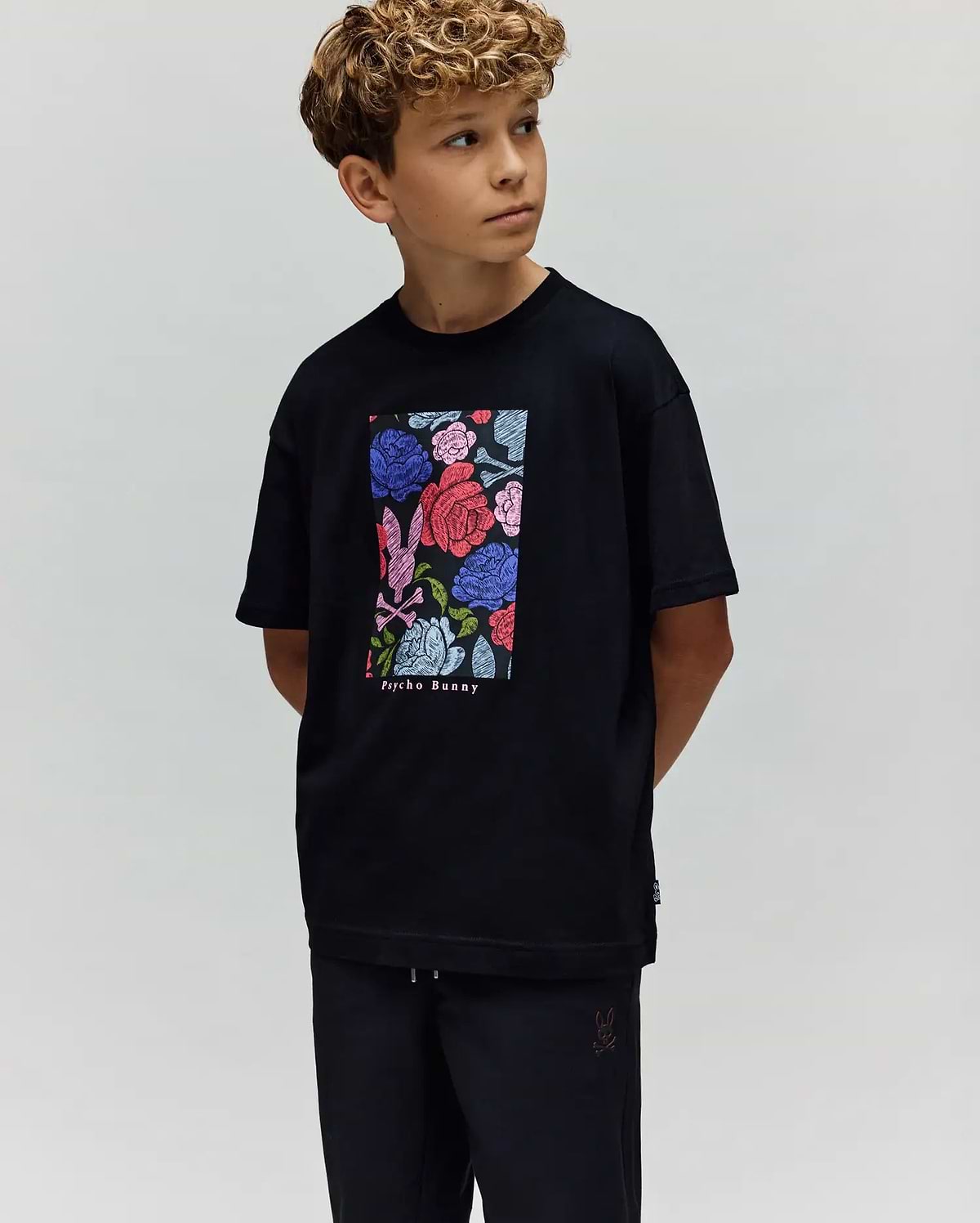 A young boy with curly hair looks to the side, wearing the Psycho Bunny KIDS ROWEN OVERSIZED TEE (KT0200093) with a colorful floral graphic and matching black pants, set against a plain light background. (1)