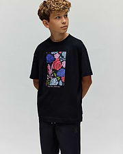 A young boy with curly hair looks to the side, wearing the Psycho Bunny KIDS ROWEN OVERSIZED TEE (KT0200093) with a colorful floral graphic and matching black pants, set against a plain light background. (1)