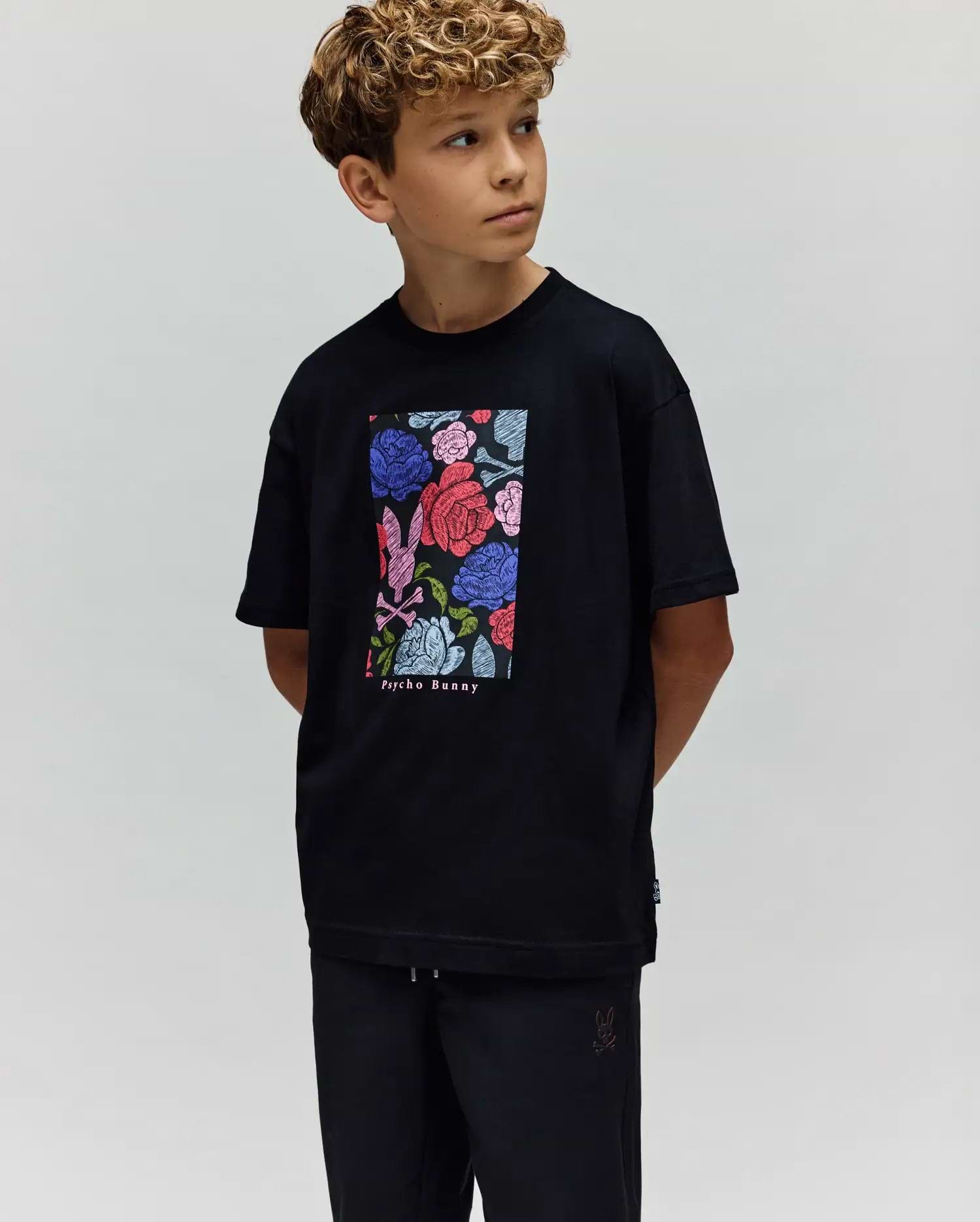 A young boy with curly hair looks to the side, wearing the Psycho Bunny KIDS ROWEN OVERSIZED TEE (KT0200093) with a colorful floral graphic and matching black pants, set against a plain light background. (1)