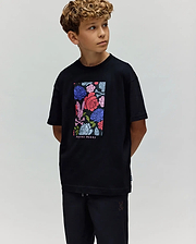 A young boy with curly hair looks to the side, wearing the Psycho Bunny KIDS ROWEN OVERSIZED TEE (KT0200093) with a colorful floral graphic and matching black pants, set against a plain light background. (1)