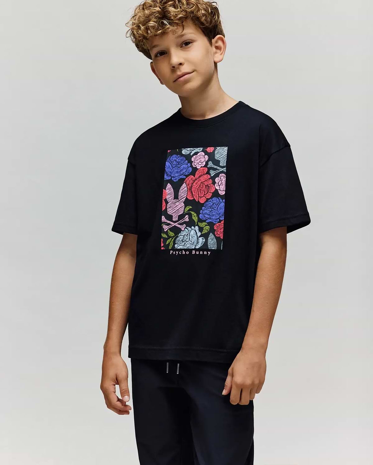 A boy with curly hair wears the Psycho Bunny KIDS ROWEN OVERSIZED TEE (KT0200093), a relaxed-fit shirt with a vibrant floral graphic and brand text, paired with black pants, standing against a plain light background. (3)