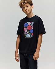 A boy with curly hair wears the Psycho Bunny KIDS ROWEN OVERSIZED TEE (KT0200093), a relaxed-fit shirt with a vibrant floral graphic and brand text, paired with black pants, standing against a plain light background. (3)