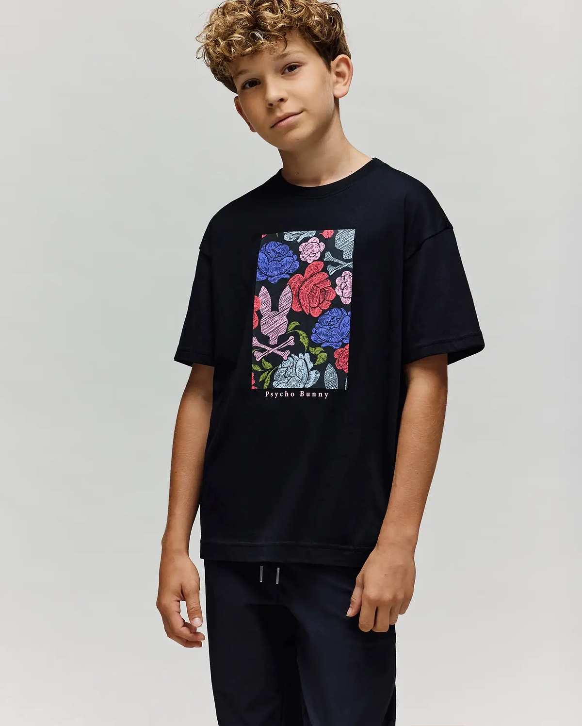 A boy with curly hair wears the Psycho Bunny KIDS ROWEN OVERSIZED TEE (KT0200093), a relaxed-fit shirt with a vibrant floral graphic and brand text, paired with black pants, standing against a plain light background. (3)