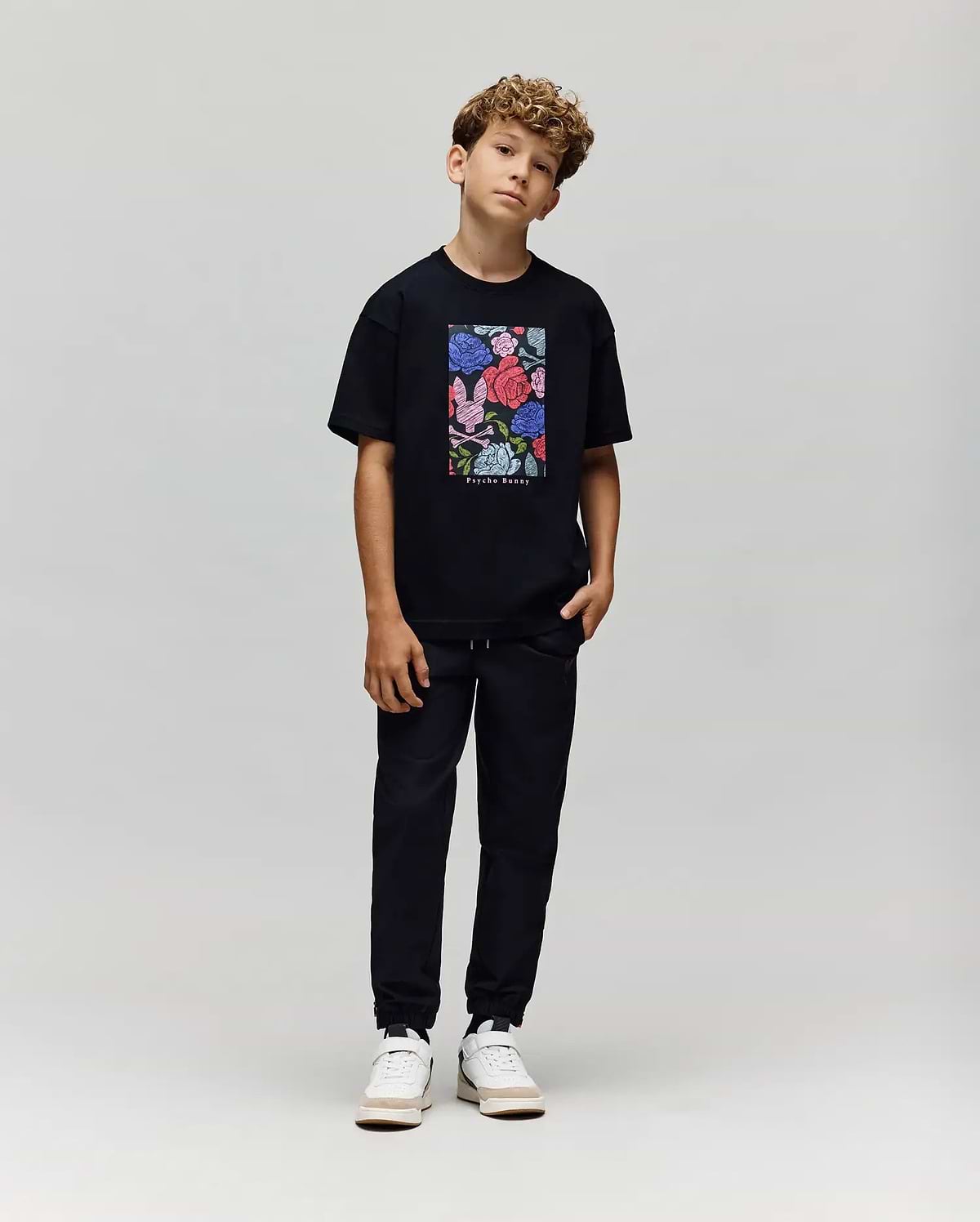A boy with curly hair stands against a plain background, wearing the Psycho Bunny KIDS ROWEN OVERSIZED TEE (KT0200093) with a colorful floral graphic, paired with black pants and white sneakers. He poses casually with his head slightly tilted. (4)