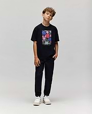 A boy with curly hair stands against a plain background, wearing the Psycho Bunny KIDS ROWEN OVERSIZED TEE (KT0200093) with a colorful floral graphic, paired with black pants and white sneakers. He poses casually with his head slightly tilted. (4)