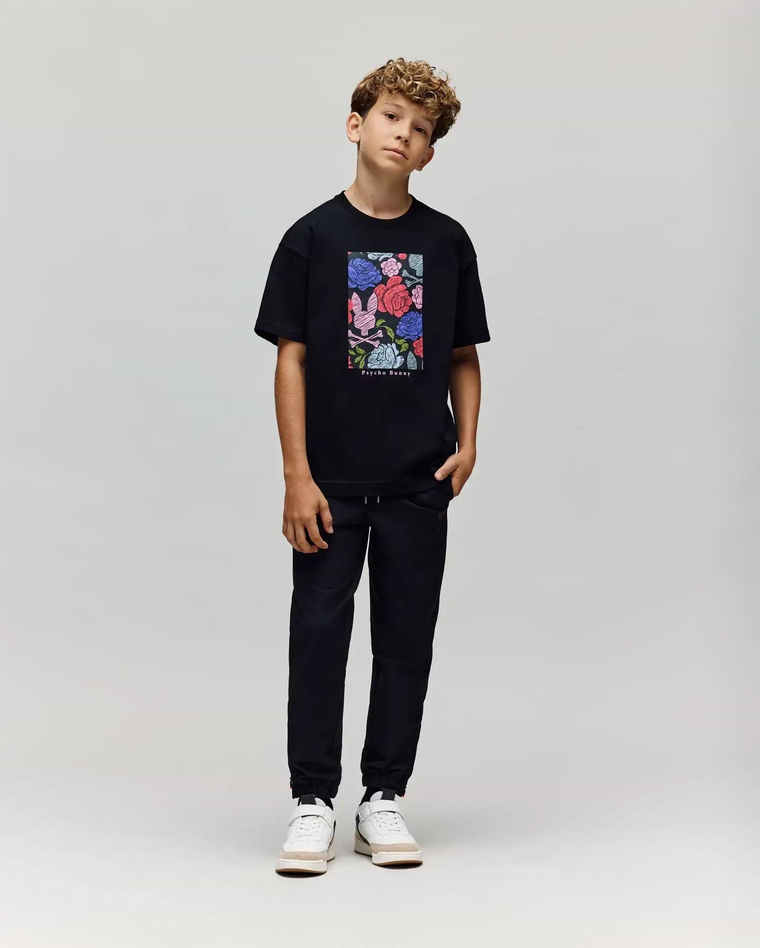 A boy with curly hair stands against a plain background, wearing the Psycho Bunny KIDS ROWEN OVERSIZED TEE (KT0200093) with a colorful floral graphic, paired with black pants and white sneakers. He poses casually with his head slightly tilted. (4)