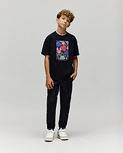 A boy with curly hair stands against a plain background, wearing the Psycho Bunny KIDS ROWEN OVERSIZED TEE (KT0200093) with a colorful floral graphic, paired with black pants and white sneakers. He poses casually with his head slightly tilted. (4)