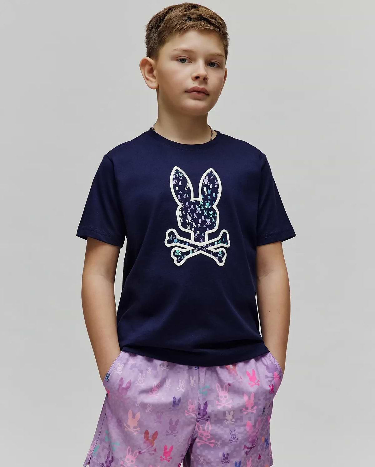Kids Nev Bunny Graphic Tee. Navy blue classic fit jersey t-shirt featuring a large center chest Psycho Bunny skull logo, filled with a vibrant multi-color mini bunny skull pattern, with lavender all-over print shorts. (2)