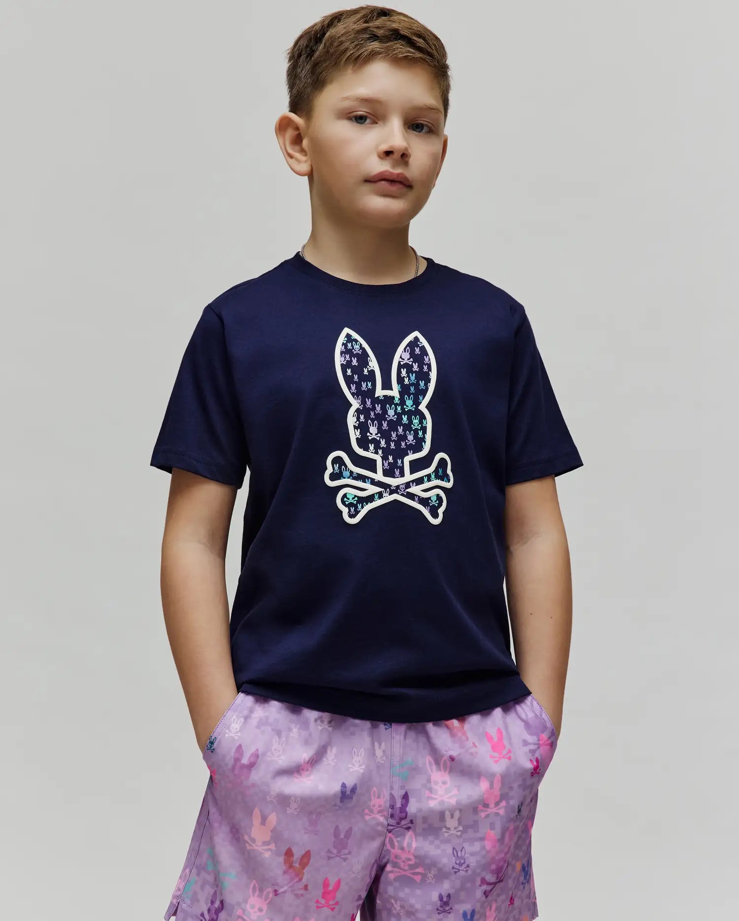 Kids Nev Bunny Graphic Tee. Navy blue classic fit jersey t-shirt featuring a large center chest Psycho Bunny skull logo, filled with a vibrant multi-color mini bunny skull pattern, with lavender all-over print shorts. (2)