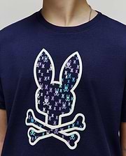 Kids Nev Bunny Graphic Tee. Navy jersey knit, classic fit. Features a large central white-outlined Psycho Bunny skull logo filled with small repeating light blue and purple bunny skulls. (3)