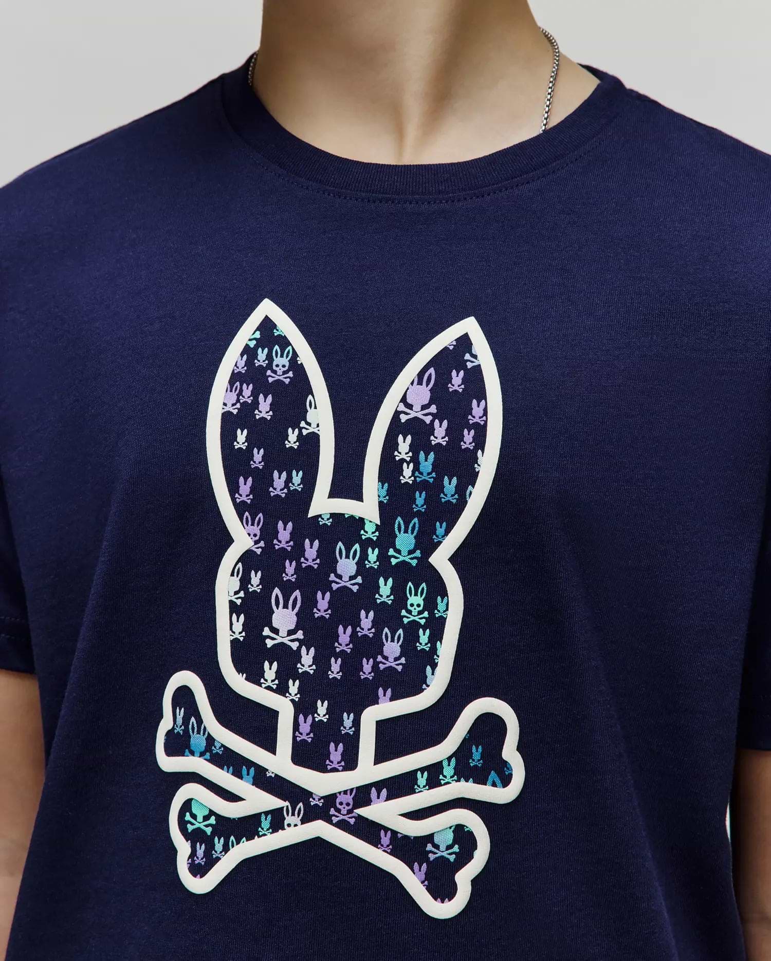Kids Nev Bunny Graphic Tee. Navy jersey knit, classic fit. Features a large central white-outlined Psycho Bunny skull logo filled with small repeating light blue and purple bunny skulls. (3)