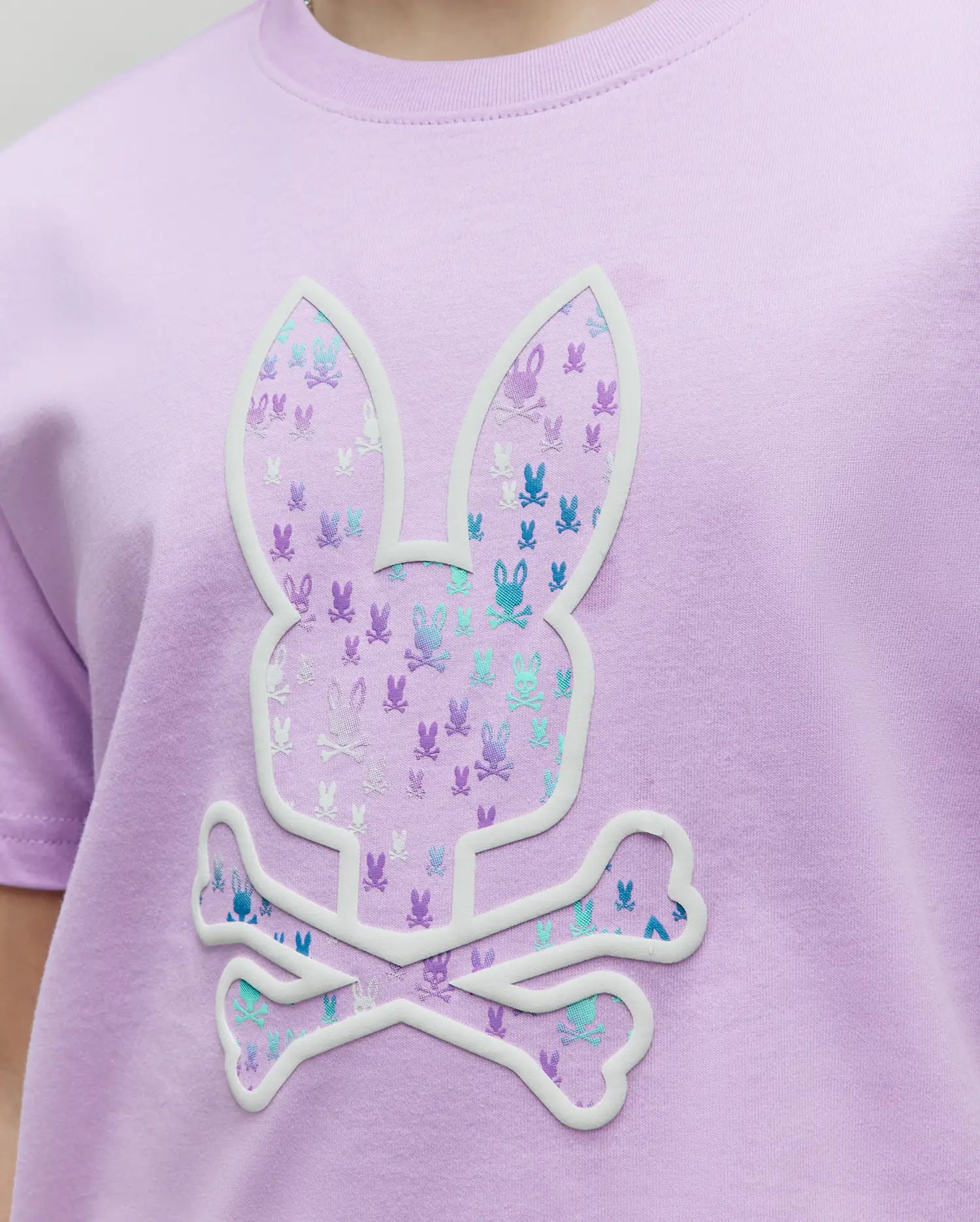 Kids Nev Bunny Graphic Tee in lavender jersey knit, featuring a large central Psycho Bunny skull logo with a raised white outline, filled with a repeating pattern of smaller multi-colored bunny skull logos. (3)