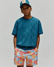 Kids Dustin Oversized Tee in deep teal, featuring an all-over embossed checkerboard pattern with Psycho Bunny logos. Worn with multi-color pixelated patchwork shorts and a teal trucker cap with a tan Psycho Bunny logo patch.