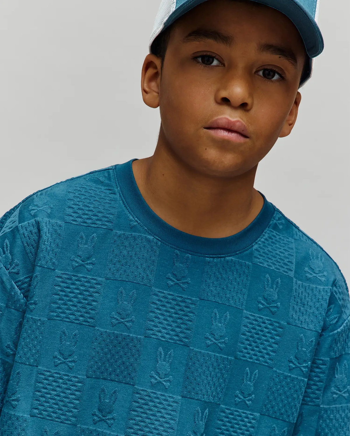 Kids Dustin Oversized Tee in teal, featuring an all-over embossed checkerboard pattern with alternating Psycho Bunny skull logos and textured squares, crafted from a soft, oversized knit fabric.