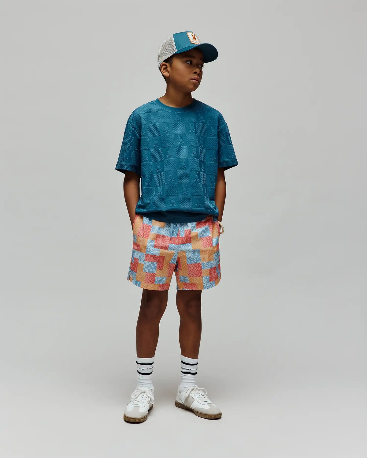 Kids Dustin Oversized Tee in teal, featuring an all-over embossed checkerboard pattern with integrated Psycho Bunny skull logos, a relaxed fit, and textured jersey fabric. Paired with multi-color patchwork shorts.