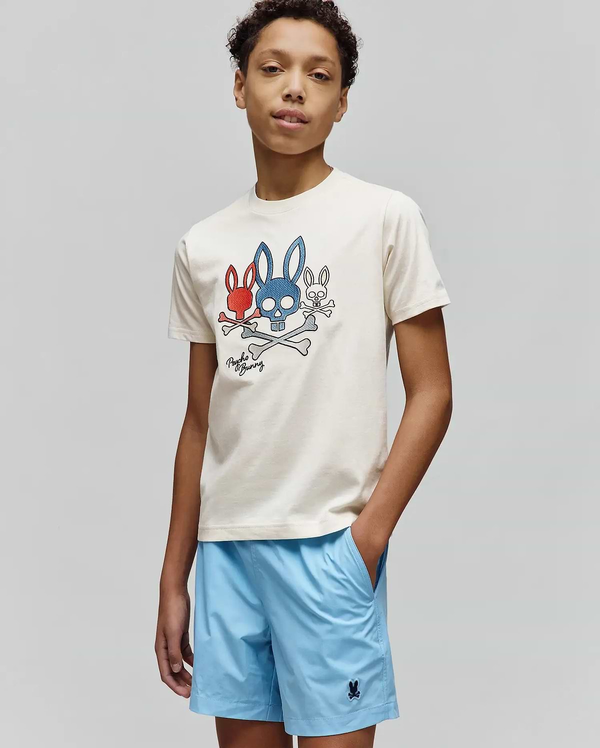 Cream Kids Oscar Graphic Tee, classic fit jersey, with a bold red, blue, white bunny skull and crossbones chest graphic. Light blue relaxed shorts feature a small embroidered Psycho Bunny logo. (4)