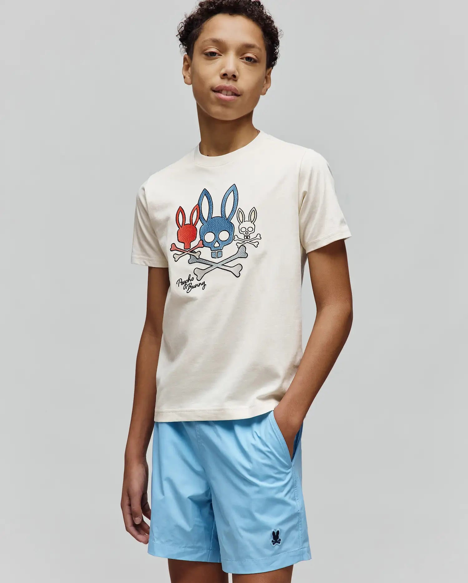 Cream Kids Oscar Graphic Tee, classic fit jersey, with a bold red, blue, white bunny skull and crossbones chest graphic. Light blue relaxed shorts feature a small embroidered Psycho Bunny logo. (4)