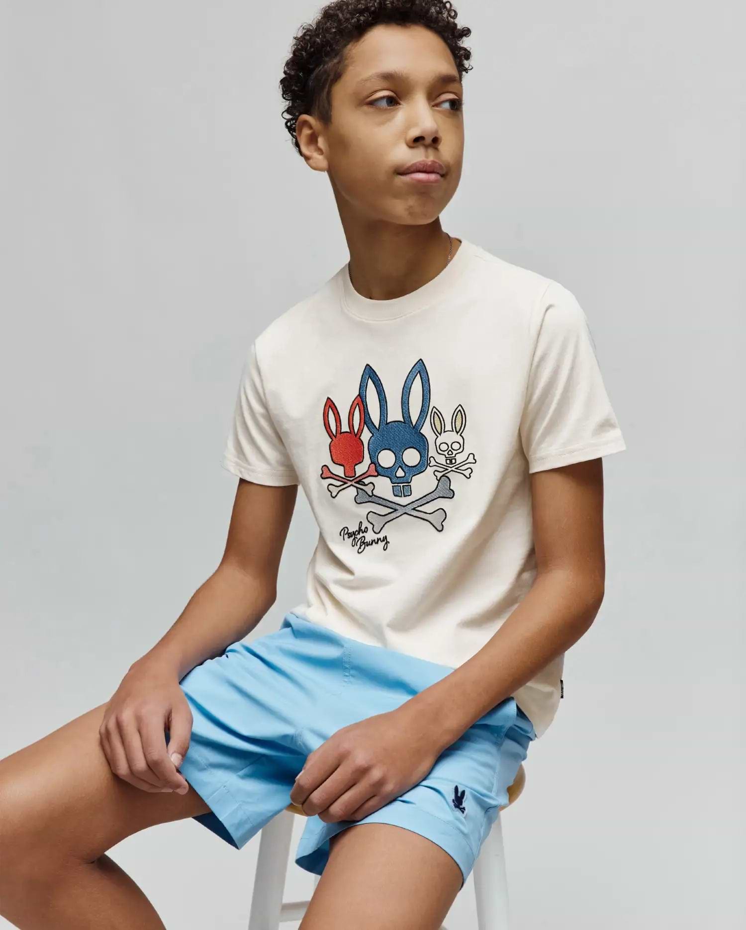 Kids Oscar Graphic Tee in cream cotton jersey, with a large central red, blue, and white Psycho Bunny skull and crossbones graphic. Light blue shorts feature a small embroidered bunny logo. (2)
