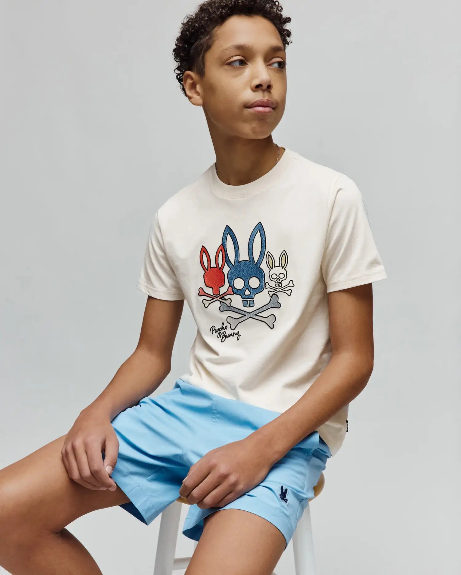 Kids Oscar Graphic Tee in cream cotton jersey, with a large central red, blue, and white Psycho Bunny skull and crossbones graphic. Light blue shorts feature a small embroidered bunny logo. (2)